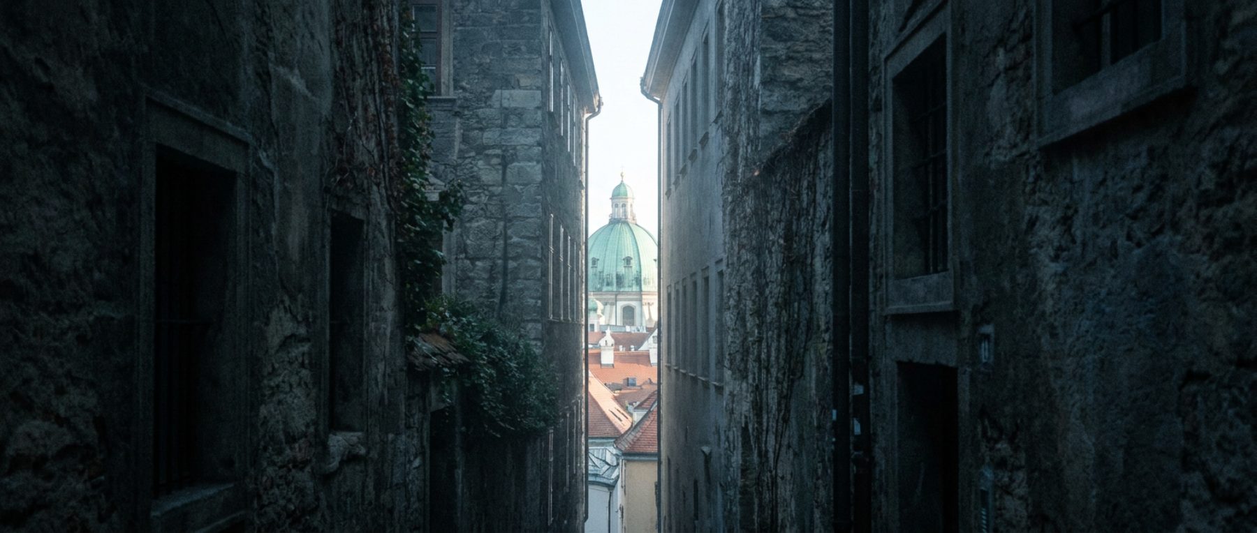 A narrow lane between dark stone buildings opening onto a bright view of rooftops and a green church dome